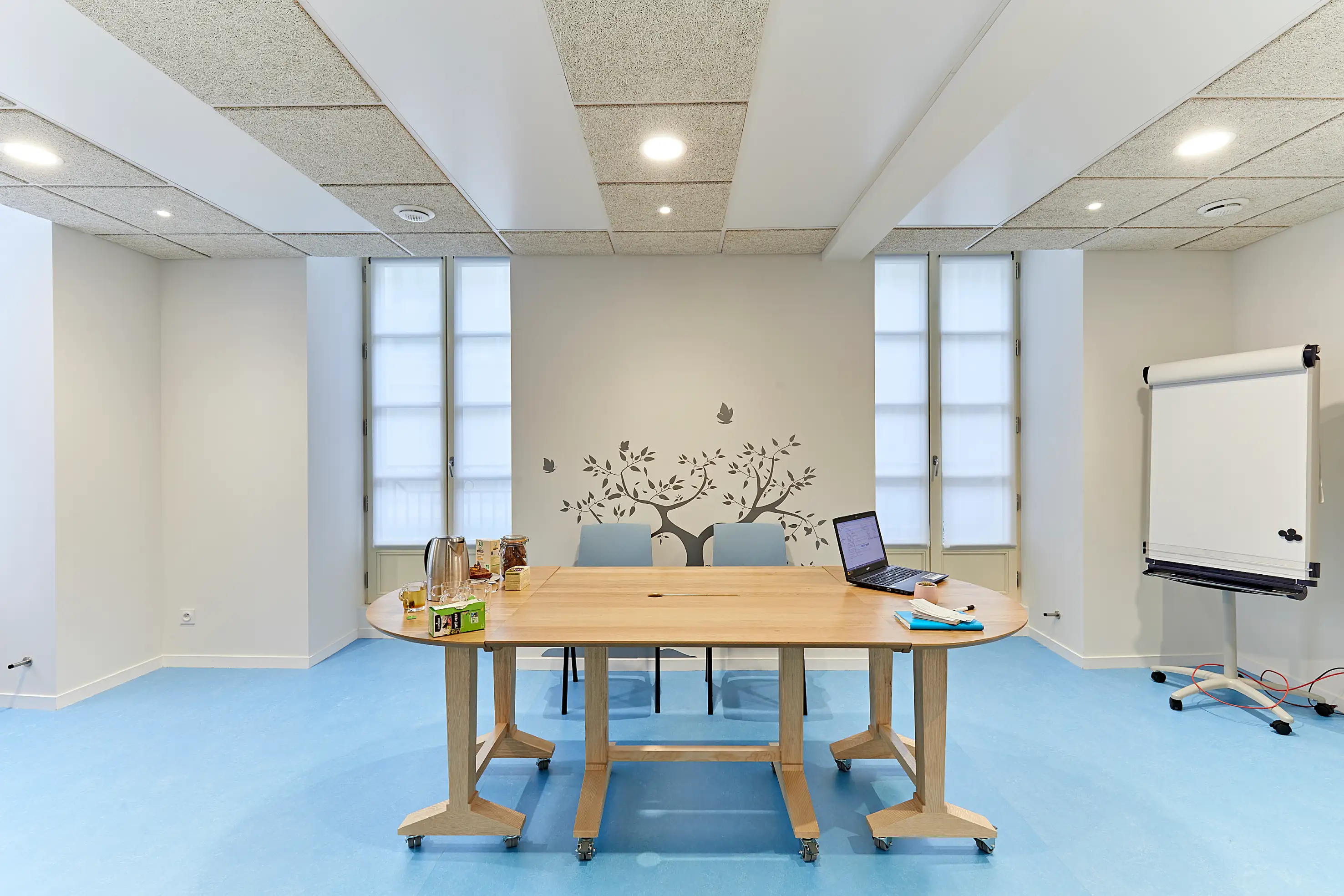 Salle de réunion lumineuse avec une table en bois clair, un sol bleu clair, un tableau blanc et de grandes fenêtres.